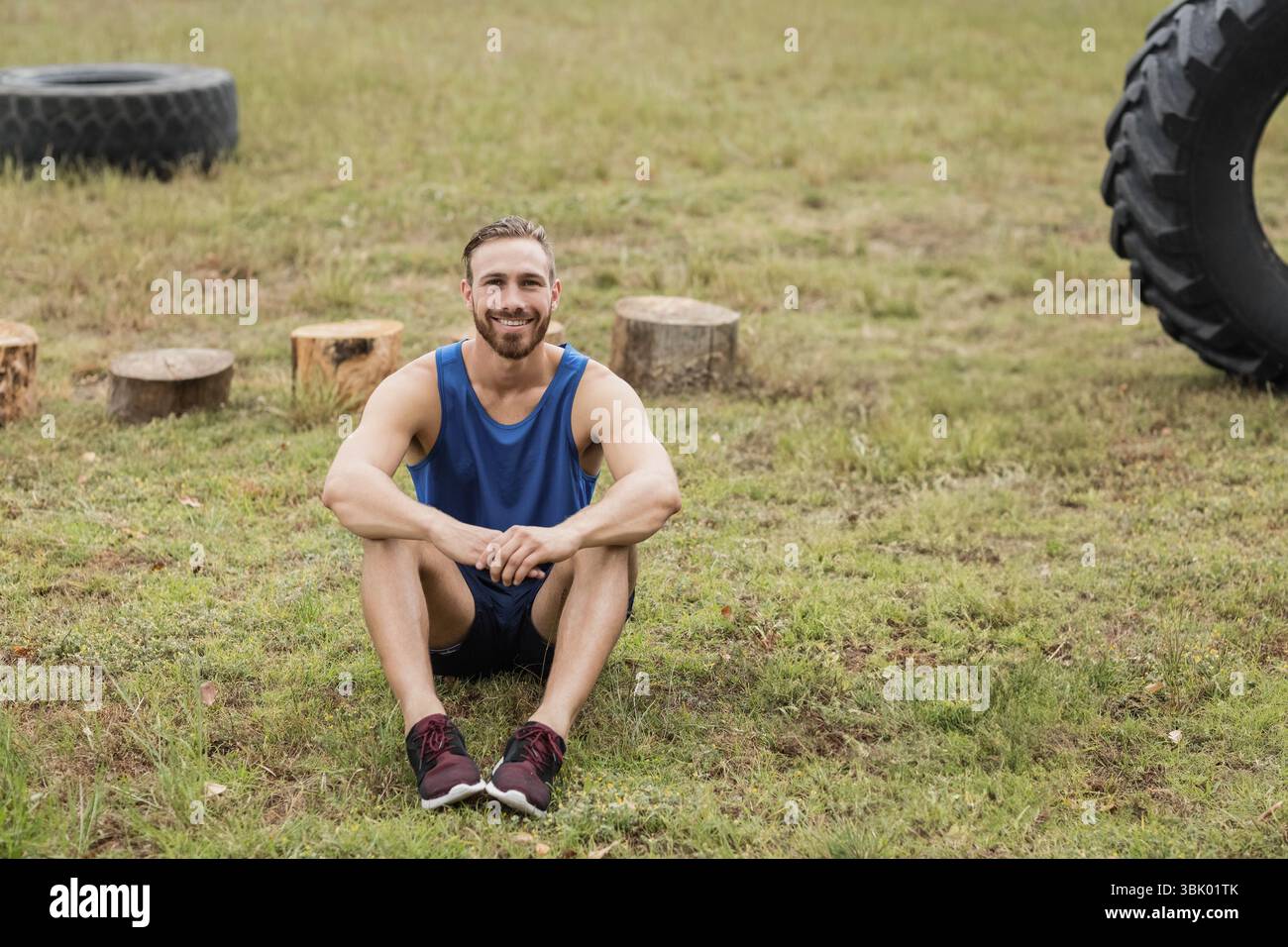 Erwachsene männliche Fitness-Enthusiast in blauem Top sitzend, lächelnd auf dem Feld mit Traktorreifen und Stümpfen Stockfoto