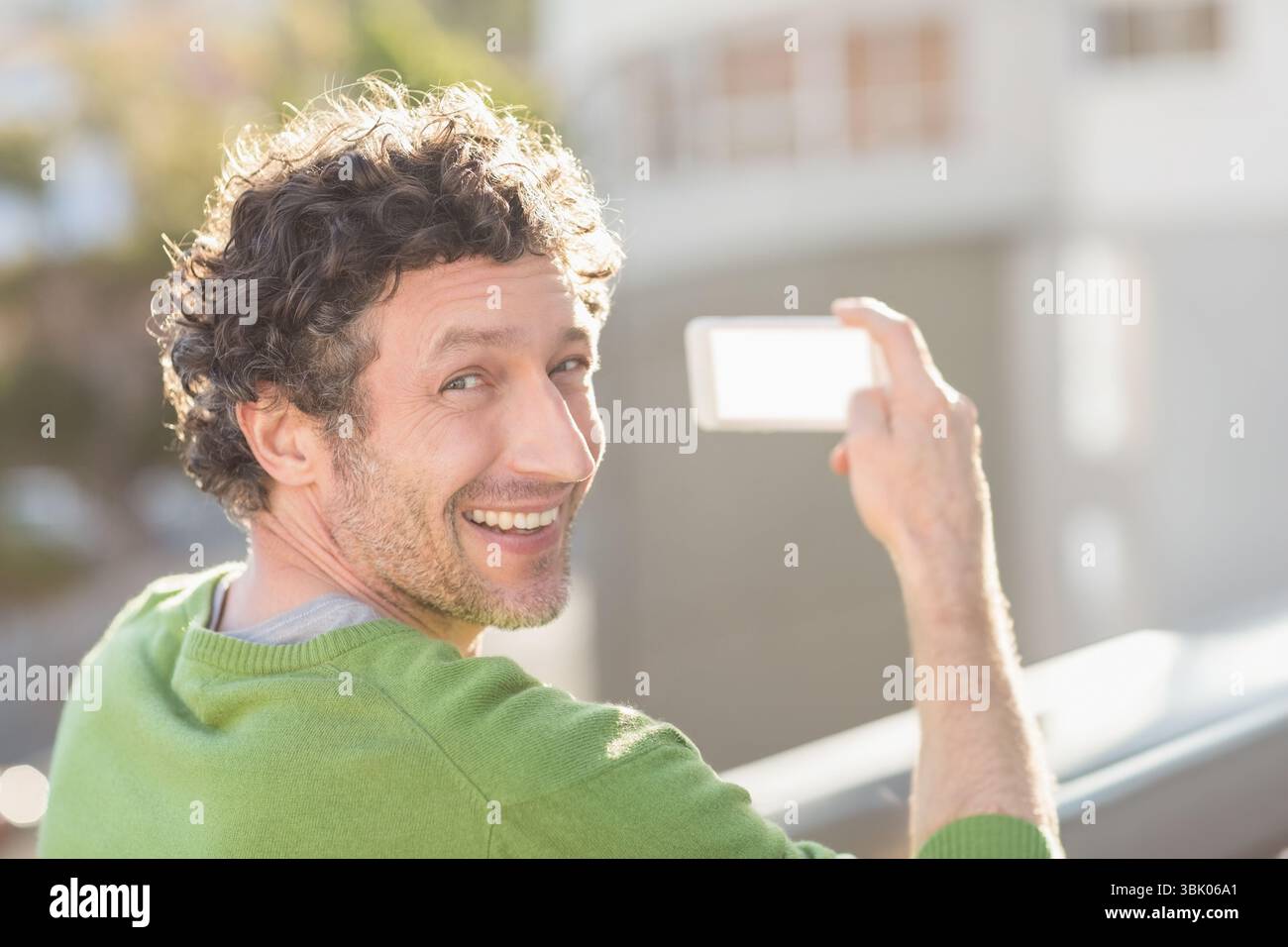Mann mit grünem Pullover, der auf einem Balkongeländer steht und ein Telefon für Selfie mit verschwommenen Fassaden hält Stockfoto