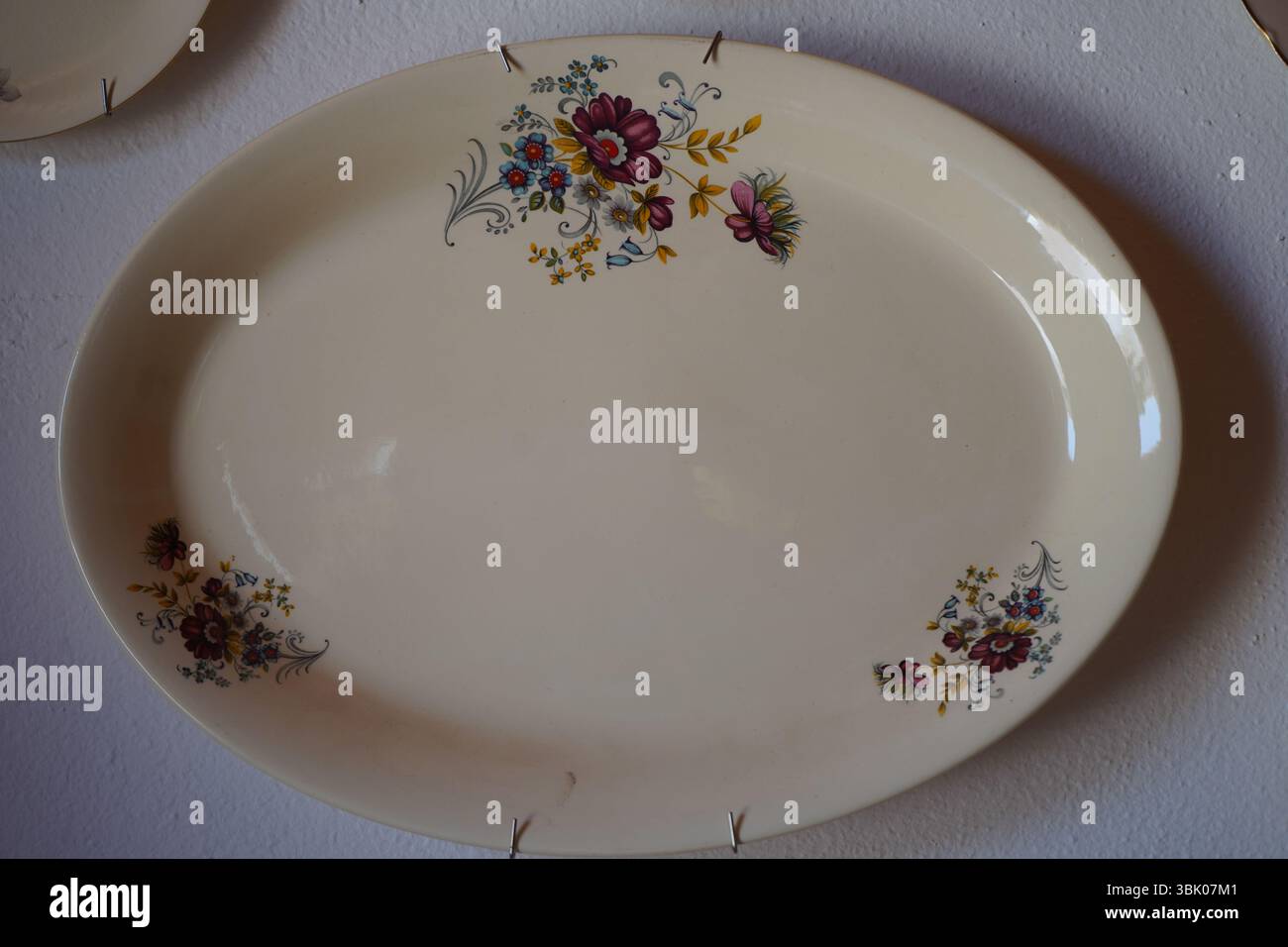 Kreta, Griechenland, 14. Juni 2025: Ovale Vintage-Keramikplatte mit Blumenmuster auf einer strukturierten weißen Wand. Stockfoto