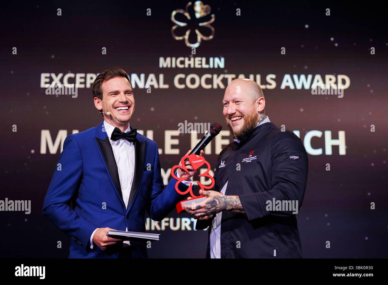 17. Juni 2025, Hessen, Frankfurt/M.: Michele Heinrich von YALDY in Frankfurt am Main (r) freut sich über seinen Cocktailpreis. Zu seiner Linken steht die Moderatorin Aljoscha Höhn. Die begehrten Michelin-Sterne werden jedes Jahr an Restaurants vergeben, die von Inspektoren für ihre gastronomische Qualität eine hohe Bewertung erhalten haben. Foto: Lando Hass/dpa Stockfoto