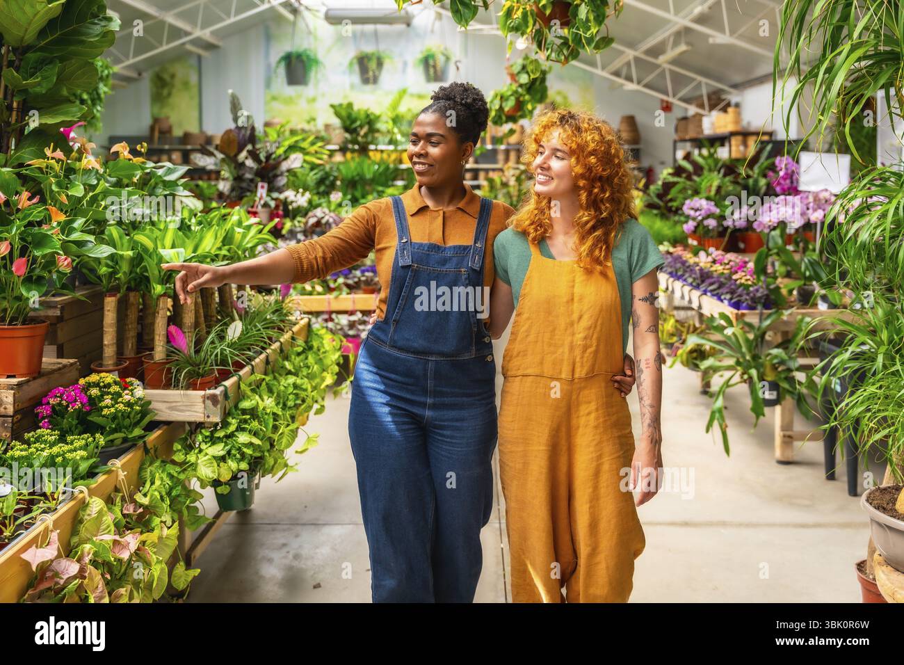 Zwei junge Frauen gehen durch ein Gartencenter, diskutieren über Pflanzen und teilen Wissen, während sie ihre Arbeit in einem lebendigen Gewächshaus genießen Stockfoto