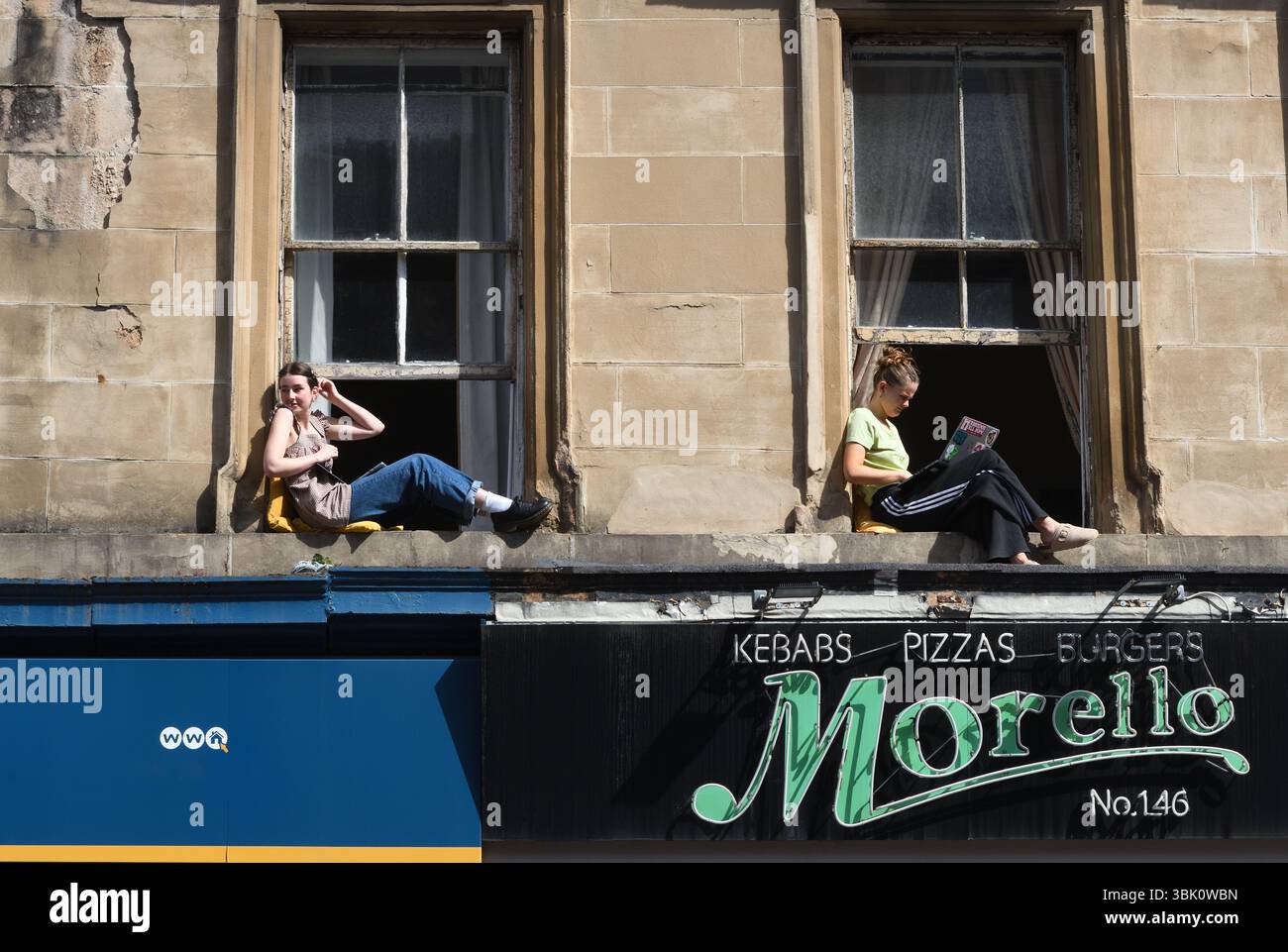 Zwei weibliche Mieter genießen die Sonne, sitzen vor den flachen Fenstern des Mietshauses in Glasgow, Schottland. Stockfoto