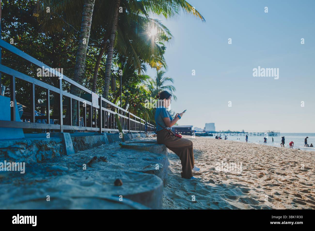 Die Frau sitzt im Schatten von Palmen, in der Ferne können andere Strandgänger die ruhige Küstenlandschaft in Balikpapan, Indon, genießen Stockfoto