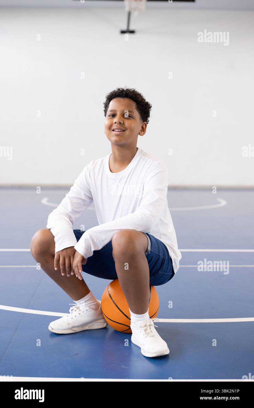 Lächelnder Junge, der im Fitnessstudio auf Basketball sitzt und sportliche Aktivitäten genießt Stockfoto