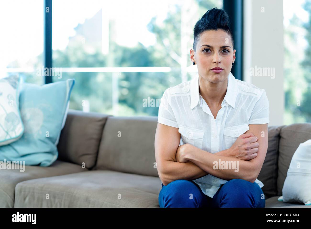 Frau aus den frühen 30er Jahren, die in der Lounge mit blauen Kissen und Fenstern auf einem taupefarbenen Sofa sitzt, Kopierraum Stockfoto
