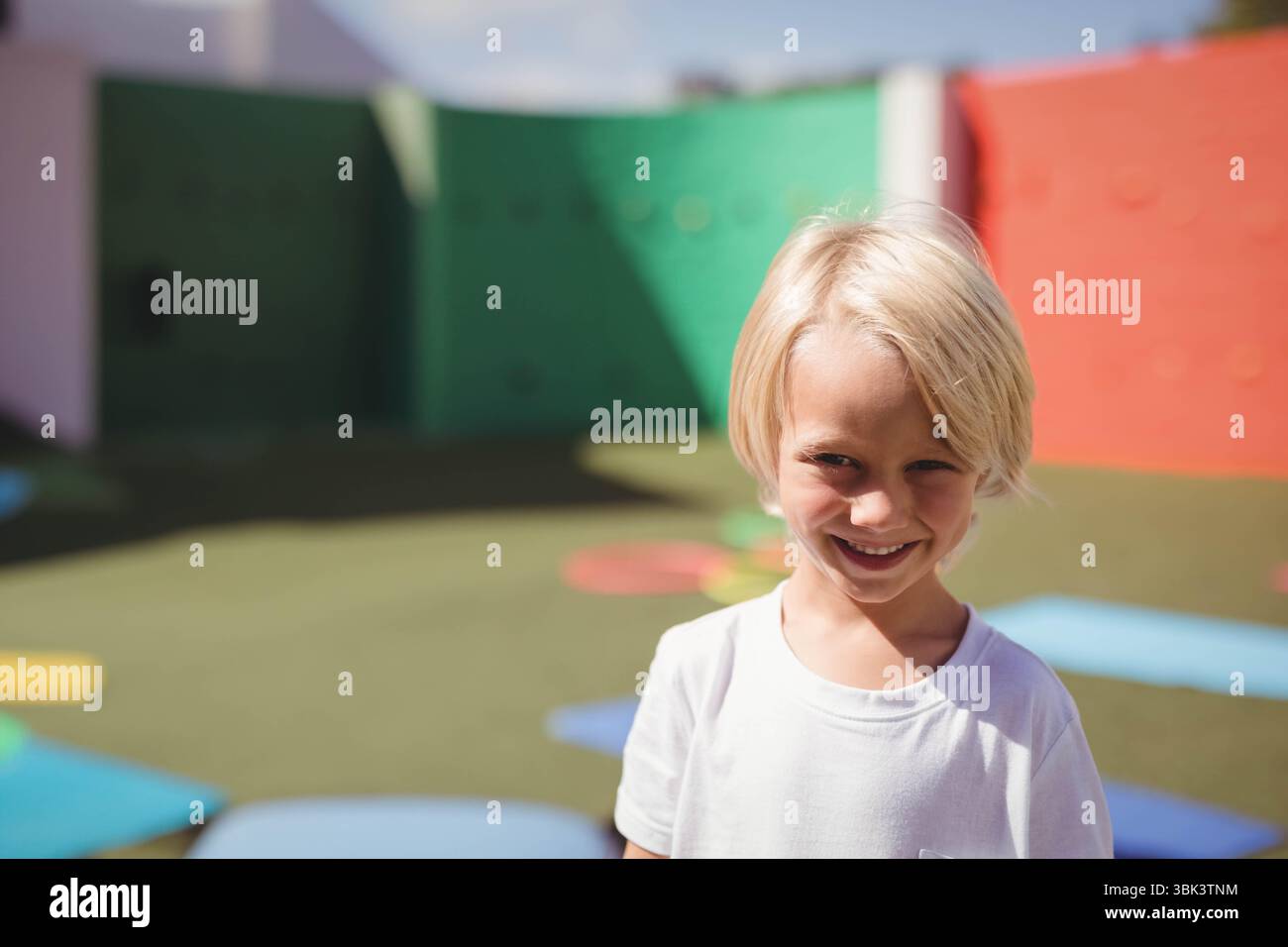 Lächelnder blonder Junge mit weißem T-Shirt auf dem Spielplatz mit Schaumstoffmatten, Klettergriffen, Kopierraum Stockfoto