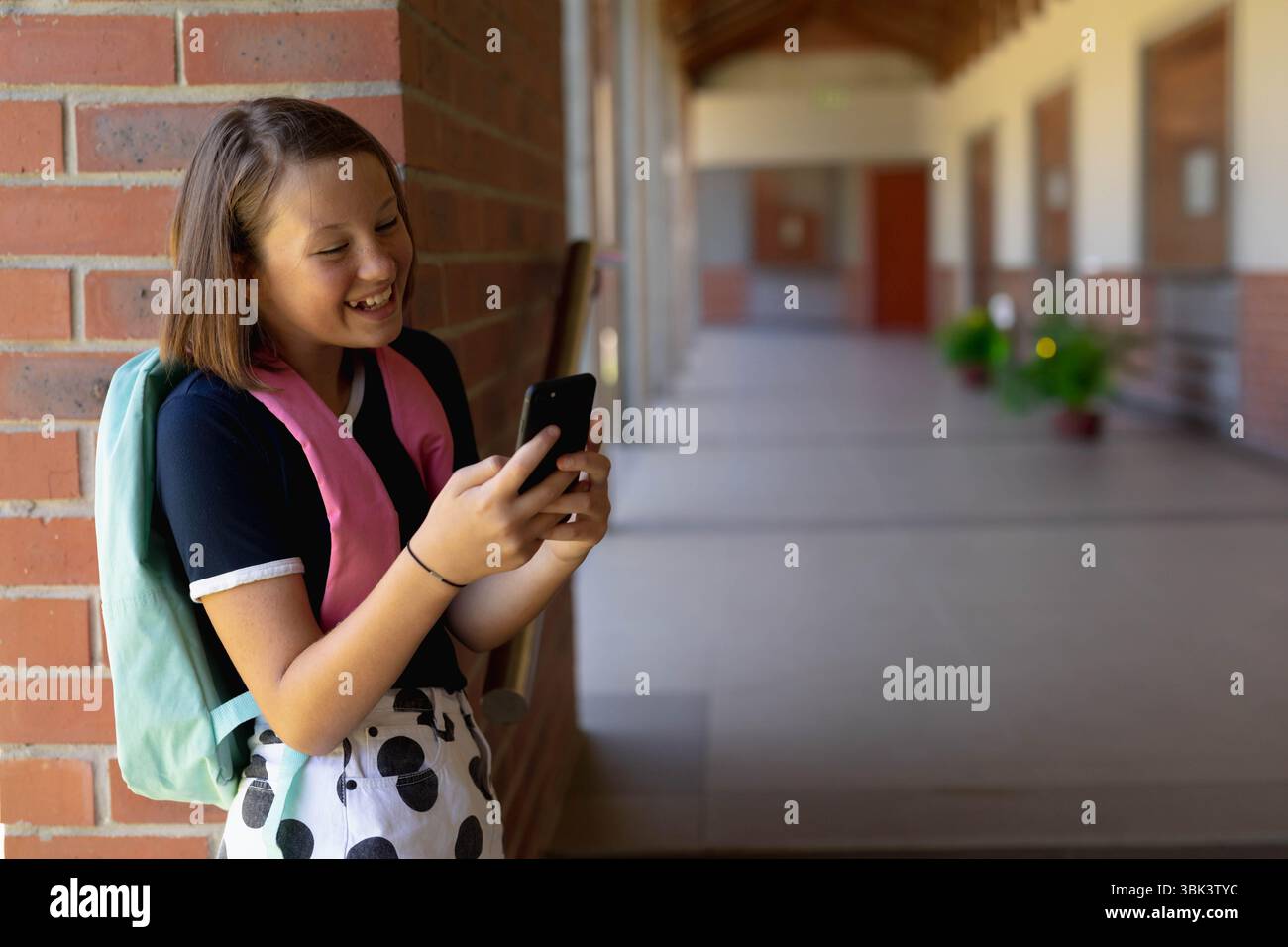 Schulmädchen lehnt sich an eine Ziegelwand mit Telefon mit Mint-Rucksack im Schulflur, Kopierraum Stockfoto