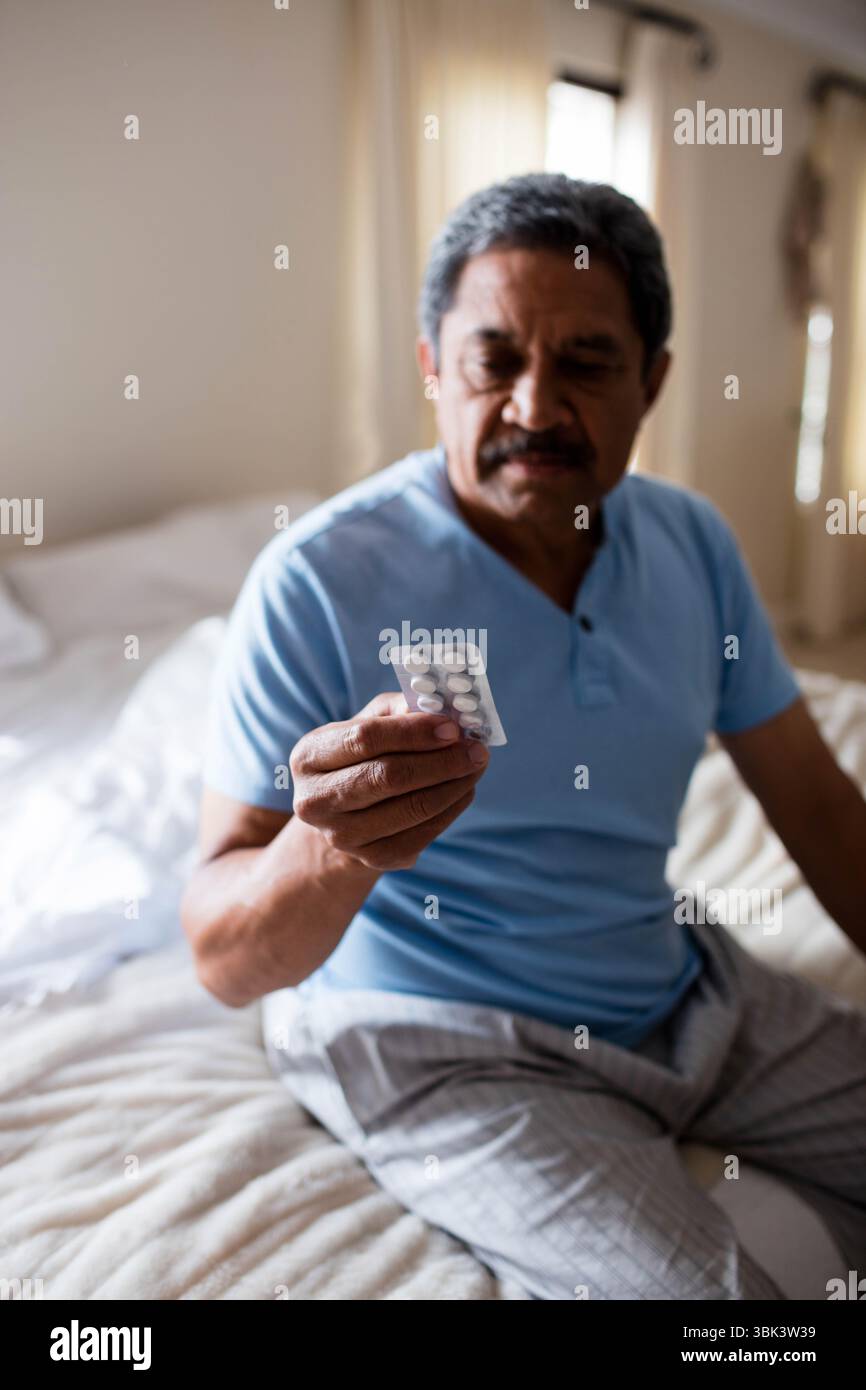 Seniorenmann mit Blisterpackung mit Tabletten, während er im Schlafzimmer mit Pyjama auf dem Bett sitzt Stockfoto