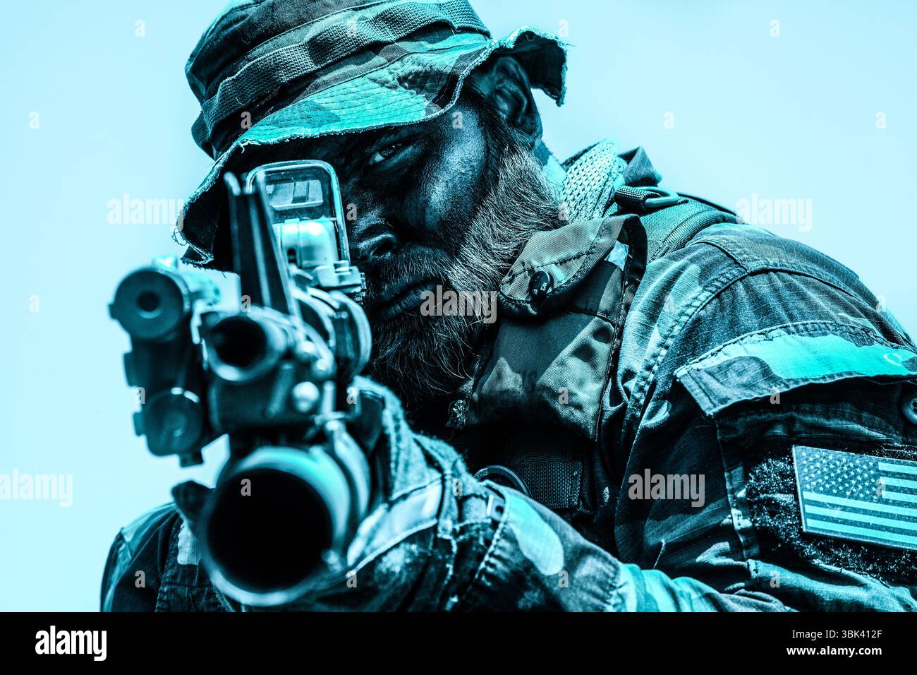 Soldat mit Tarnschutz und Helm, der ein Gewehr mit intensiver Fokussierung, ein amerikanischer Flaggenaufnäher, sichtbar auf Uniform, vor einem hellen Himmel. Stockfoto