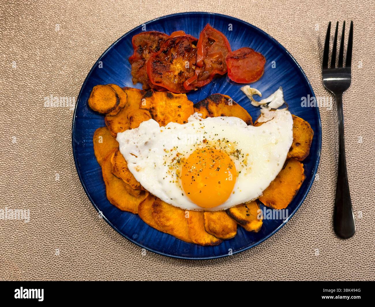 Das Frühstück besteht aus gebratenem, sonnigen Ei, Tomatenscheiben und Süßkartoffeln auf blauem Gericht in flacher Lage mit Blick über dem Zimmer Stockfoto
