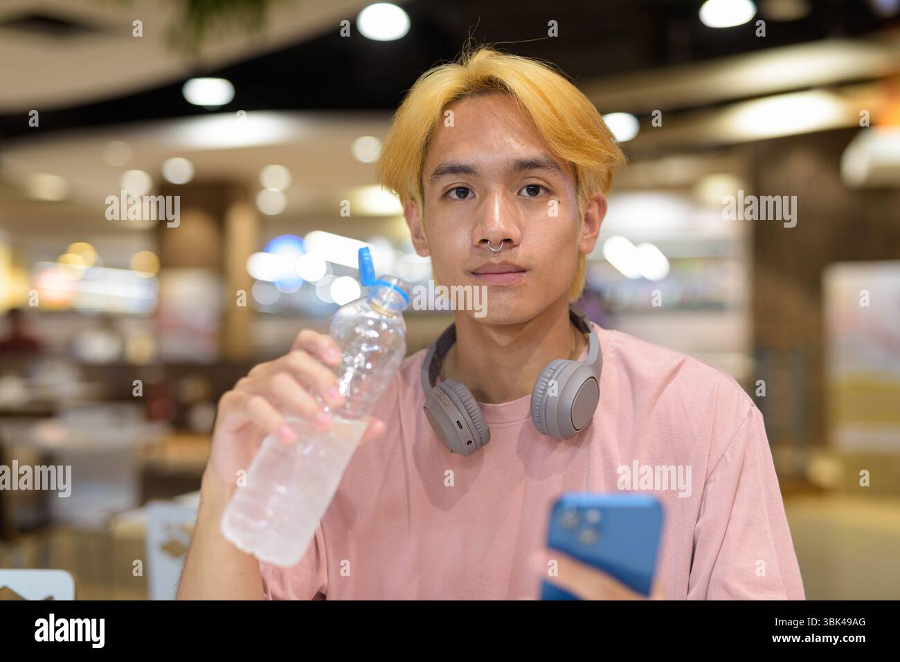 Junger Student Teenager Mann mit Kopfhörern, Trinkwasser und Handy drinnen Stockfoto