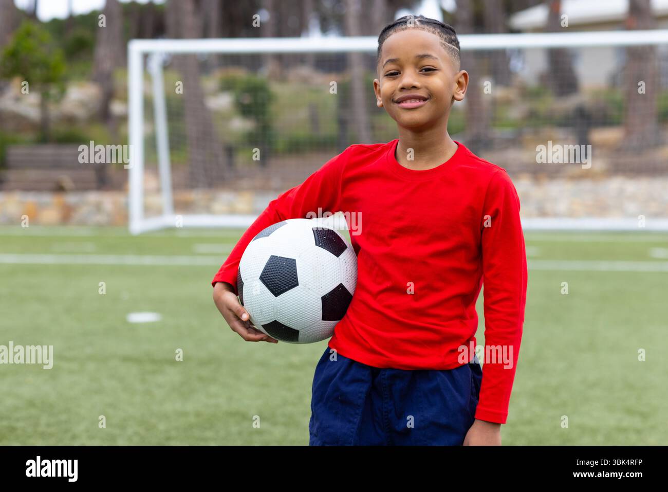 Lächelnder Junge, der Fußball auf dem Spielfeld hält und Outdoor-Aktivitäten genießt Stockfoto