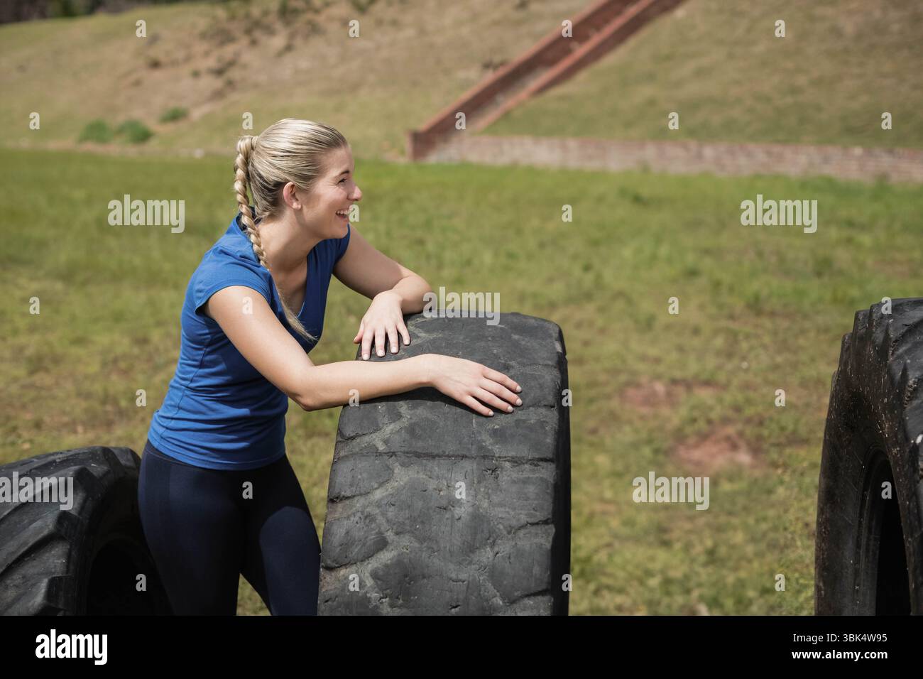 Frau, die sich auf großen Traktorreifen im grasbewachsenen Outdoor-Trainingsbereich stützt und Stärke demonstriert Stockfoto
