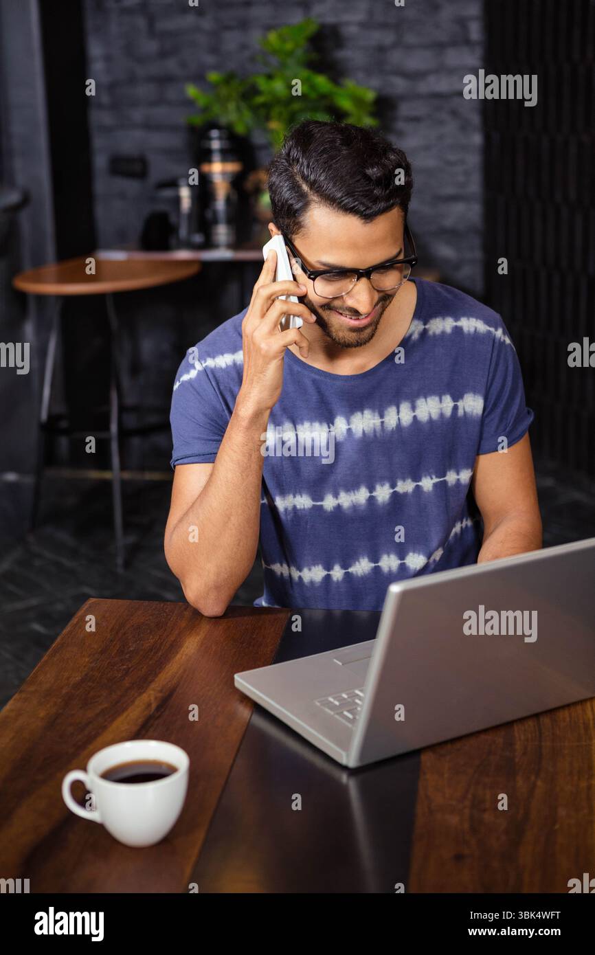 Indischer Mann mit Brille am Tisch, hält Smartphone, während er Laptop mit Kaffeetasse ansieht Stockfoto