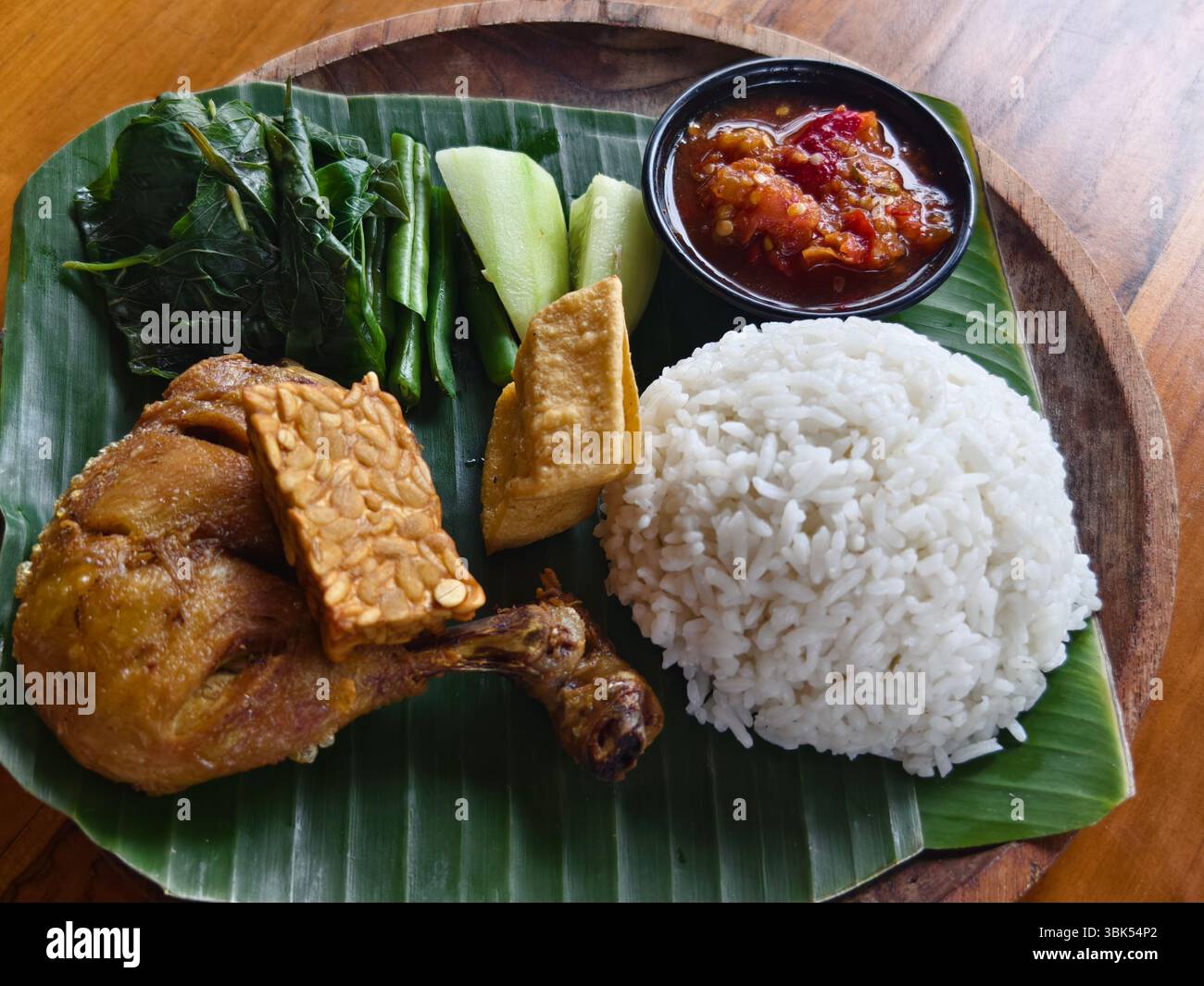 Traditionelle indonesische Mahlzeit oder Nasi Ayam Lalapan, bestehend aus gebratenem Huhn, Tempeh, Tofu, Gemüse, Sambal und gedämpftem Reis auf einer Bananenblattplatte Stockfoto