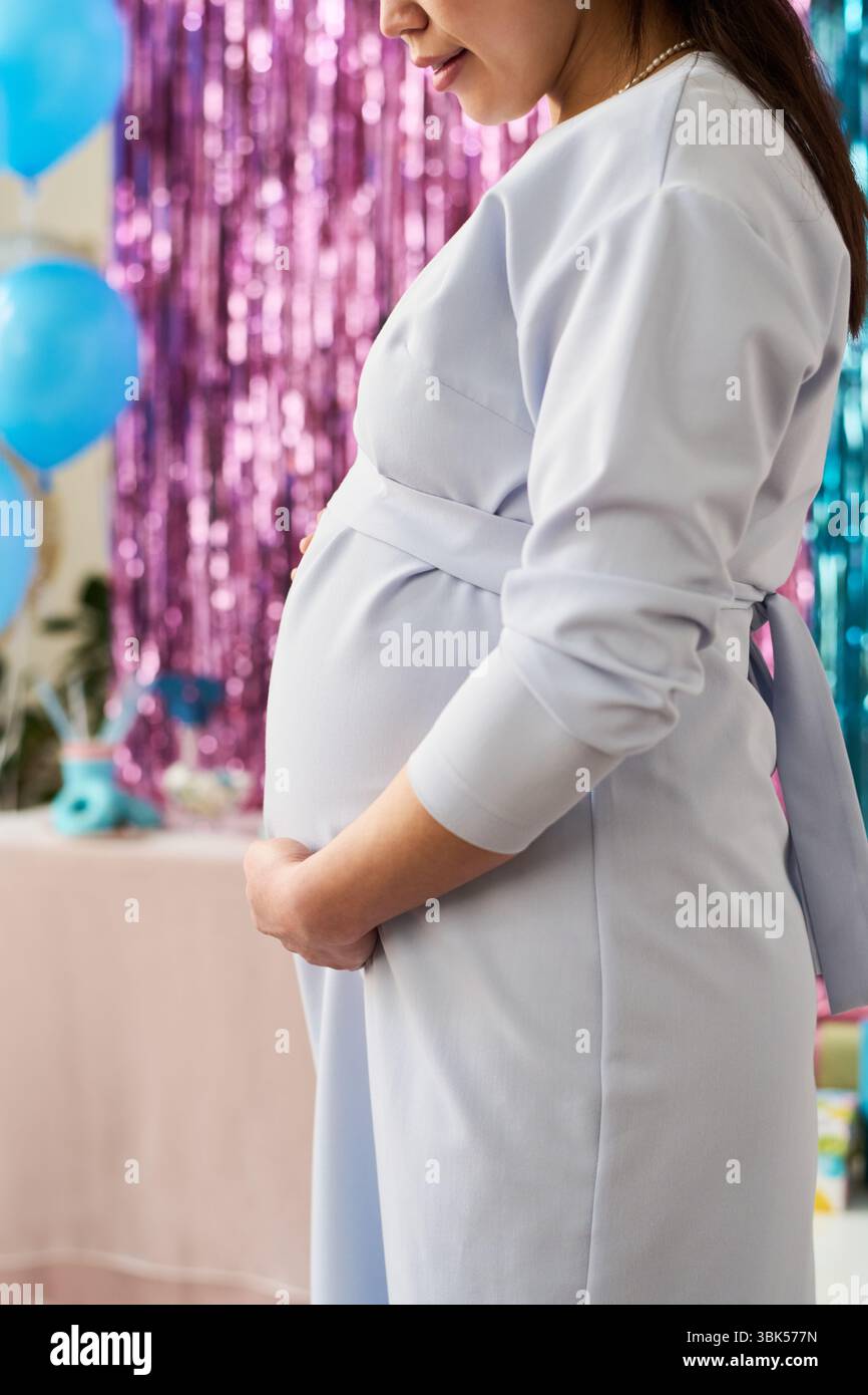 Schwangere asiatische junge Erwachsene Frau, die im Profil steht und während der Gender-Enthüllungs-Party mit bunten Ballons und Dekorationen sanft den Bauch hält Stockfoto
