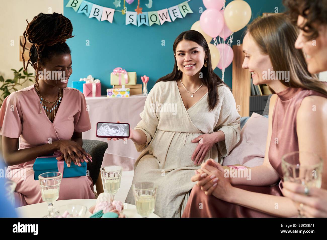 Schwangere junge Erwachsene kaukasische Frau, die mit verschiedenen Gruppen junger erwachsener Frauen sitzt und Ultraschallbilder auf dem Smartphone zeigt Stockfoto
