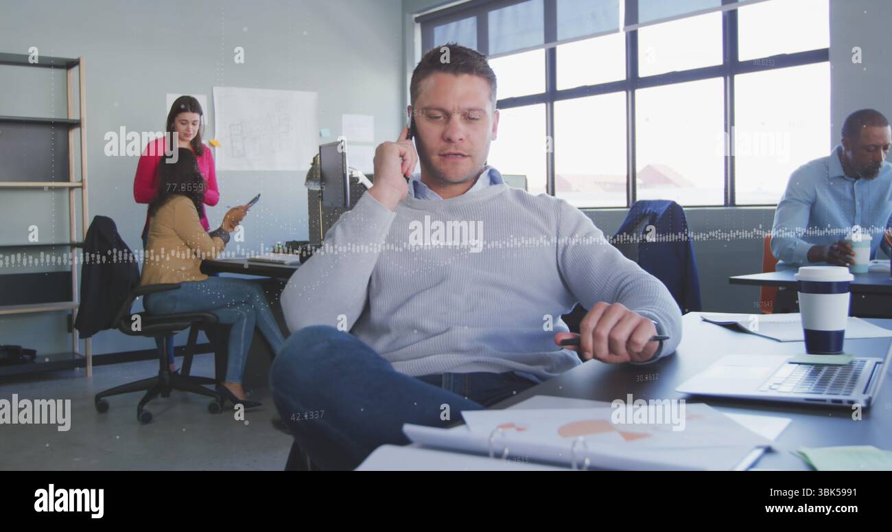 Sprechender Mann, der einen hellgrauen Pullover trägt, telefoniert am Schreibtisch, mit Laptop und Papieren Stockfoto