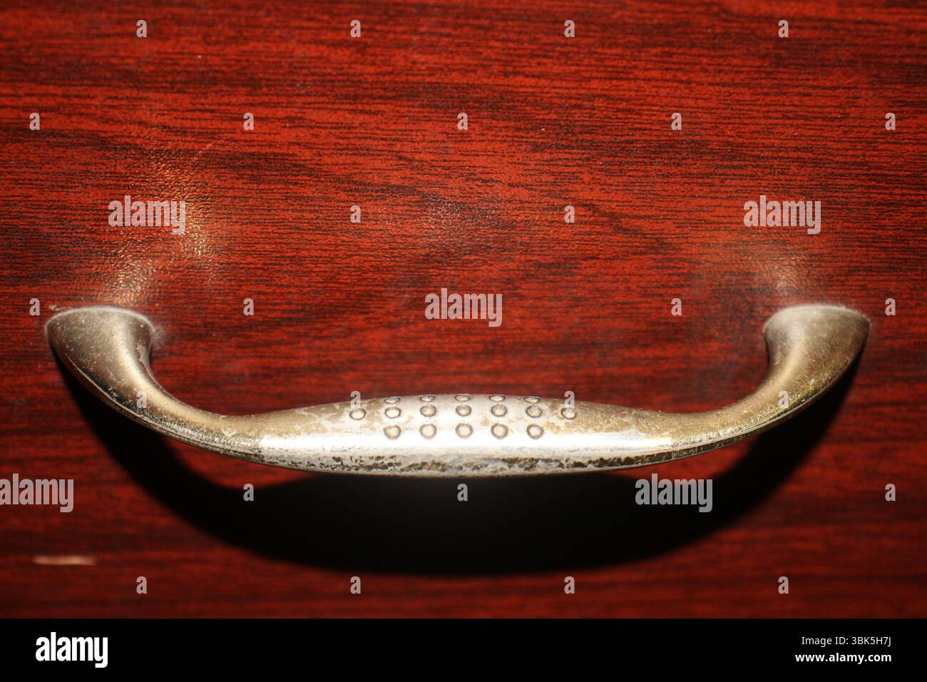 Ein Metallgriff, der an einer Holzschublade befestigt ist. Der Griff hat eine abgenutzte, leicht angetrübte Textur mit kleinen kreisförmigen Einbuchtungen in der Mitte. Stockfoto