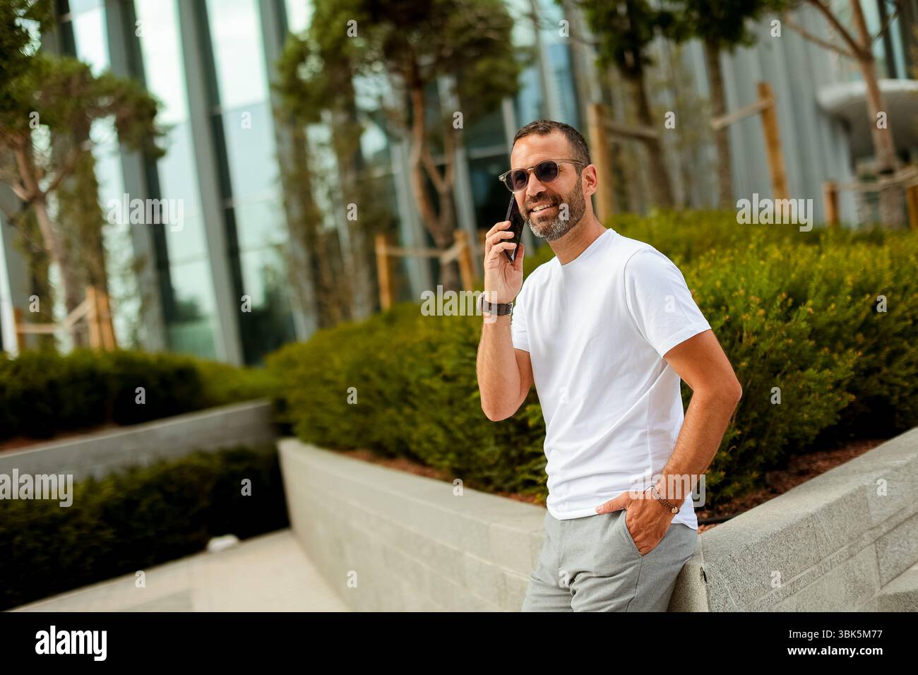 Helles Sonnenlicht strahlt ein warmes Leuchten aus, während ein lächelnder Mann in lässiger Kleidung auf seinem Smartphone spricht, während er in einem modernen grünen Raum steht. Stockfoto