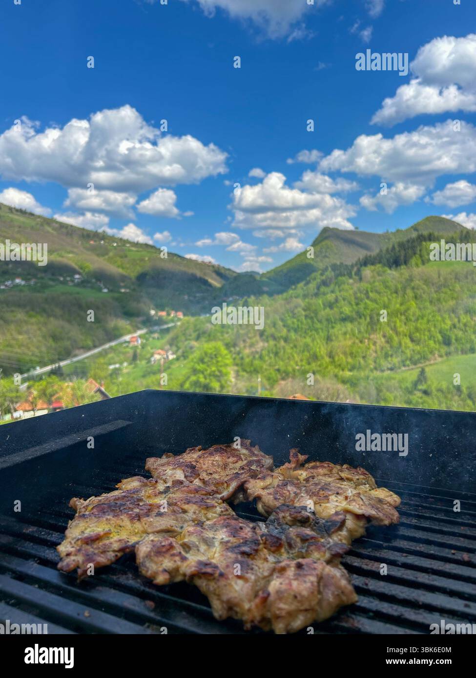 Saftiges gegrilltes Fleisch auf einem Grill mit aufsteigendem Rauch, vor einer atemberaubenden Kulisse aus grünen Bergen, blauem Himmel und flauschigen Wolken auf einer sonnigen Sonne - Smartphone-aufgenommenes Stockfoto