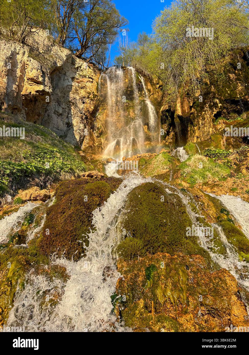 Malerischer Wasserfall, der über moosbedeckte Felsen stürzt, umgeben von üppiger Vegetation und Klippen unter einem leuchtend blauen Himmel. Ideal für Natur und Reisen - Smartphone-aufgenommenes Stockfoto
