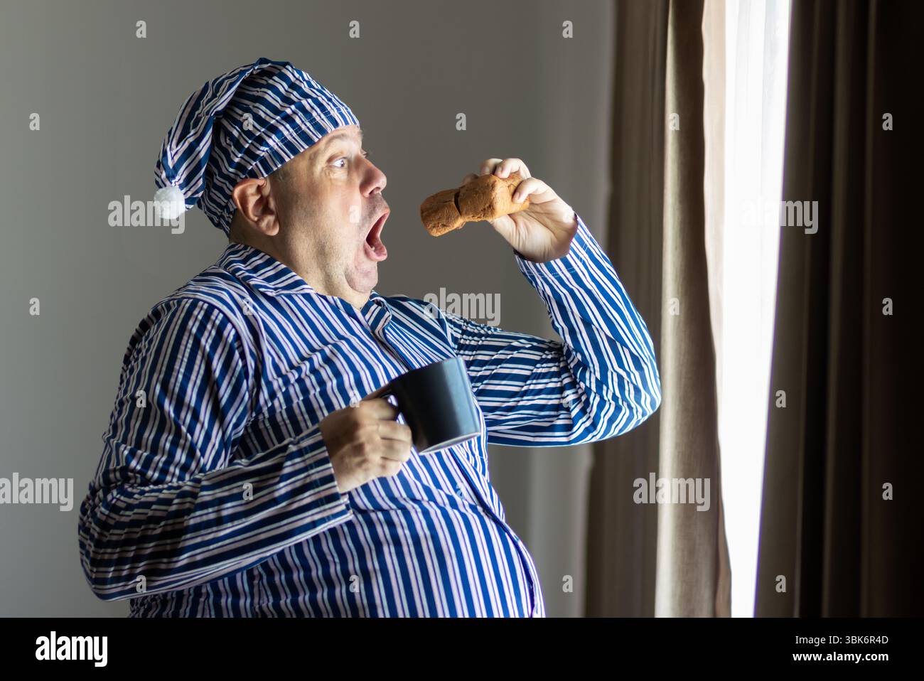 Fette Mann im Pyjama, die am Fenster frühstücken Stockfoto