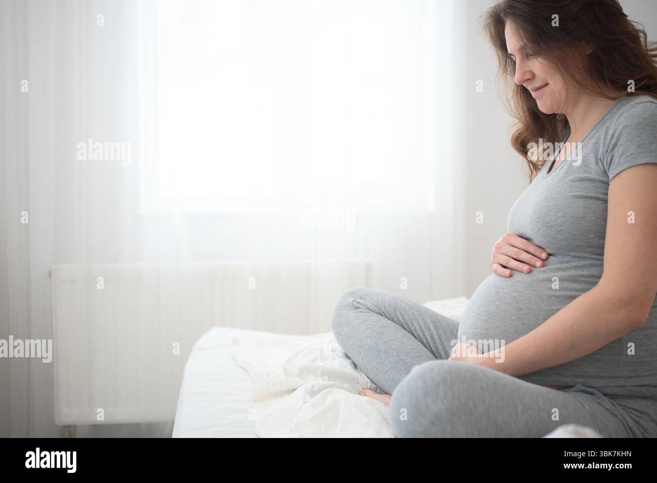 Eine heitere schwangere Frau sitzt in Lotusposition auf einem gemütlichen Bett und streichelt ihren Bauch sanft. Die warme Atmosphäre spiegelt die freudige Erwartung der Mutterschaft wider, und sanfte Beleuchtung verstärkt den emotionalen Moment. Stockfoto