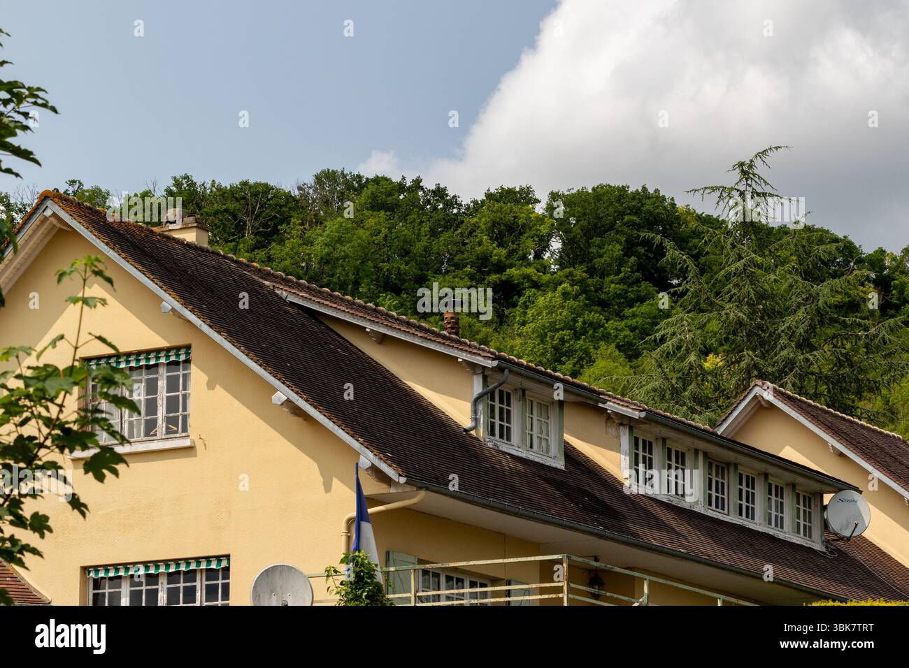 Ein braunes Gebäude mit dunklem Dach steht unter Bäumen und einem bewölkten Himmel, teilweise eingerahmt von Laub auf der linken Seite in Giverny, Frankreich. Stockfoto