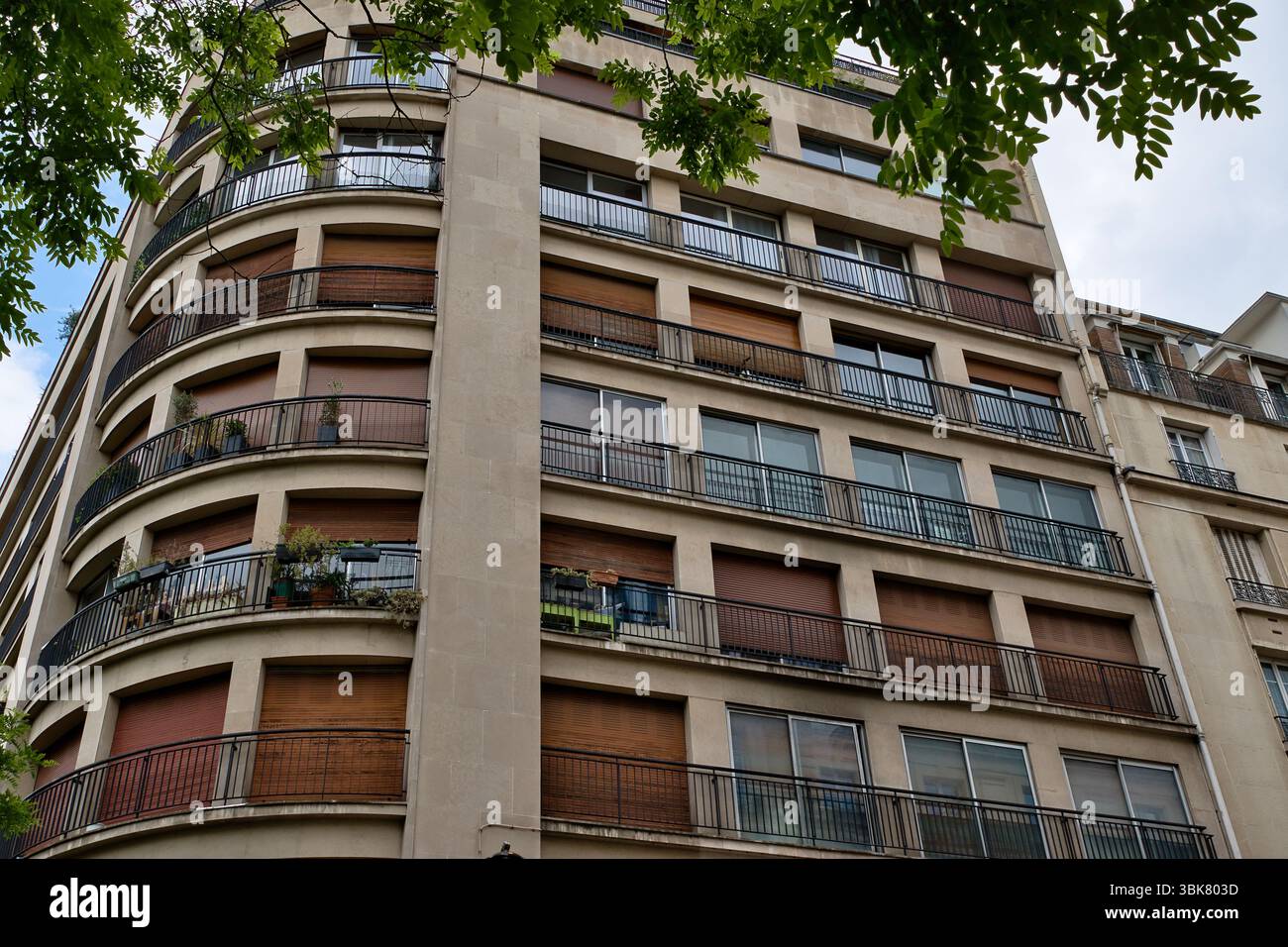 Flaches Bild eines mehrstöckigen Wohngebäudes mit Balkonen und neutralen Tönen, eingerahmt von Baumlaub an der oberen und linken Ecke. Stockfoto