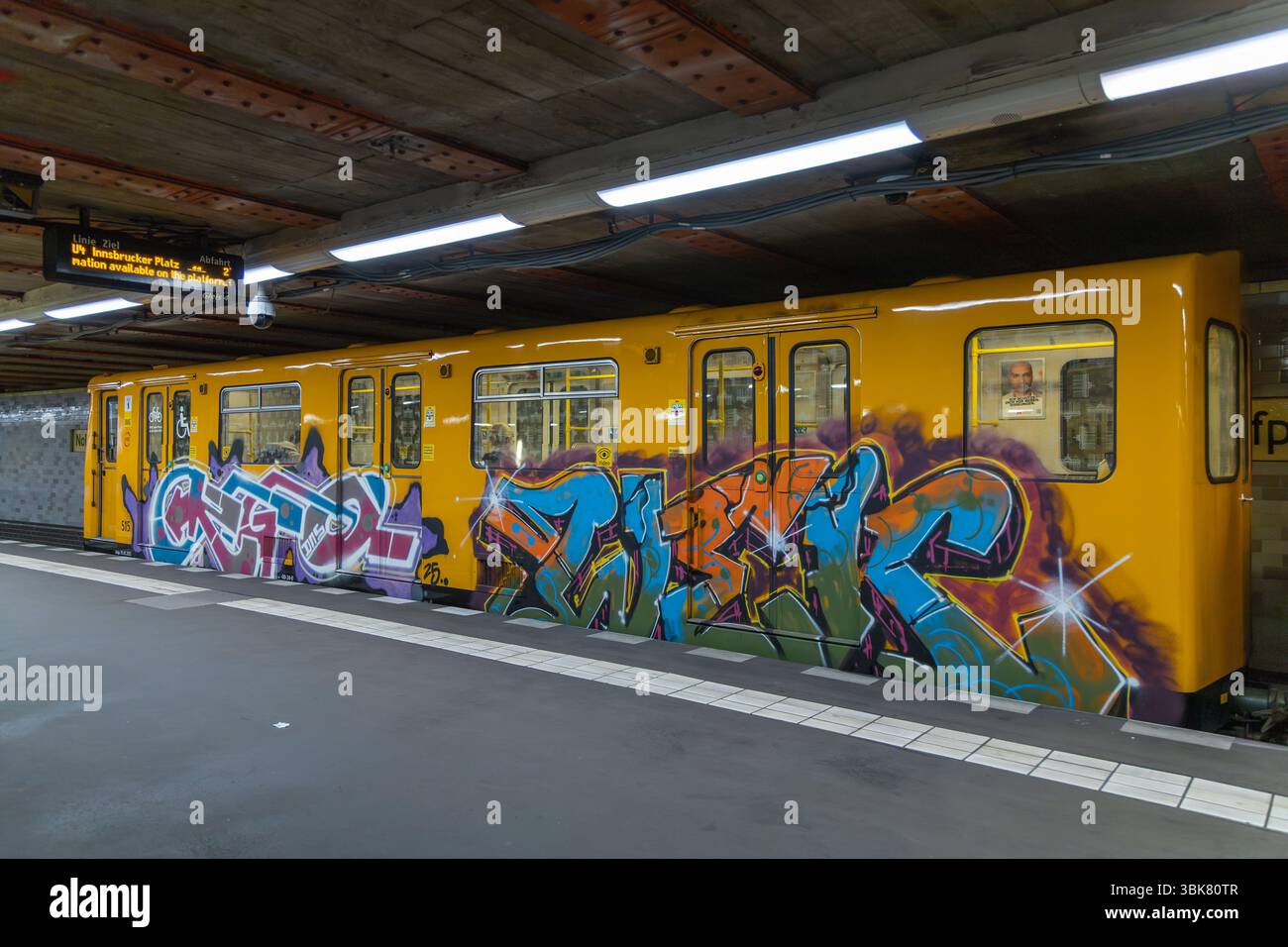 An einem Bahnsteig mit Deckenbeleuchtung steht ein gelber U-Bahn-Wagen mit lebendiger Graffiti-Kunst. Berlin, Deutschland. 28. März 2025. Stockfoto