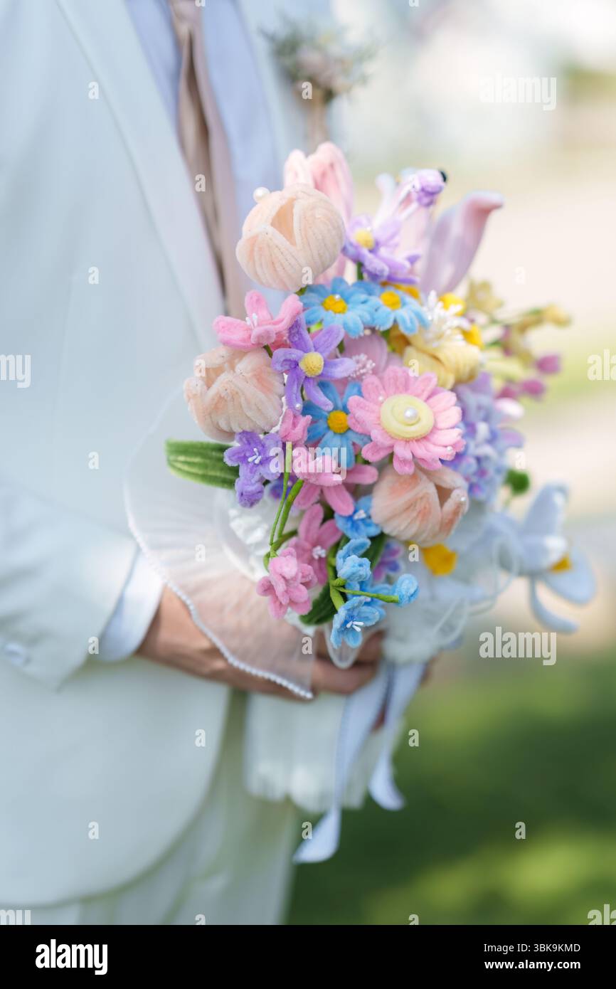 Großaufnahme des Bräutigams in weißem Anzug, der während der Hochzeitsfeier eine künstliche Blumenstraußblume in Pastellfarben hält Stockfoto