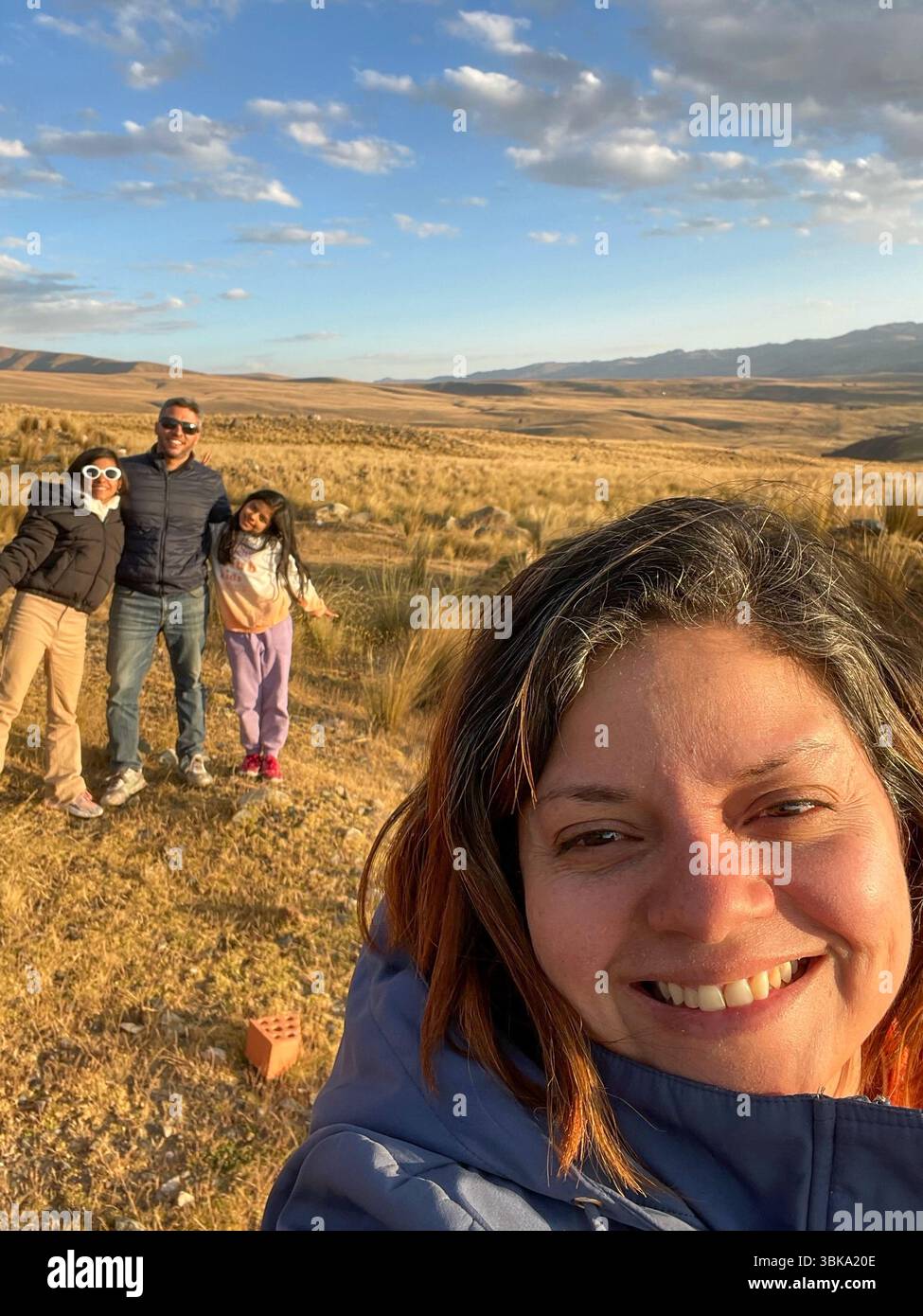 Eine Frau mit Kamera lächelt und posiert für ein Foto mit ihrer Familie. Die Familie steht auf einem Feld mit einem wunderschönen blauen Himmel über ihnen Stockfoto