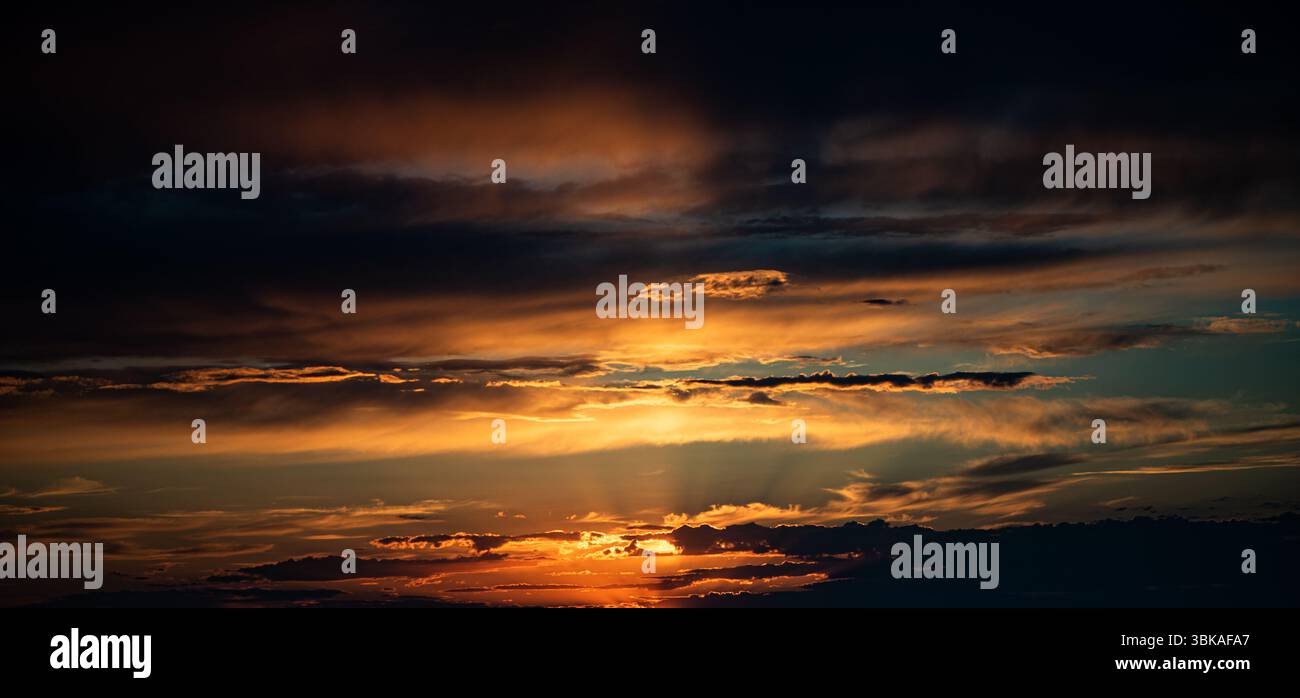 Das Foto fängt einen Sonnenuntergang mit leuchtend orangefarbenen und goldenen Sonnenstrahlen ein, die durch überlagerte Wolken brechen. Der Horizont ist dunkel und der Himmel ist voller dra Stockfoto