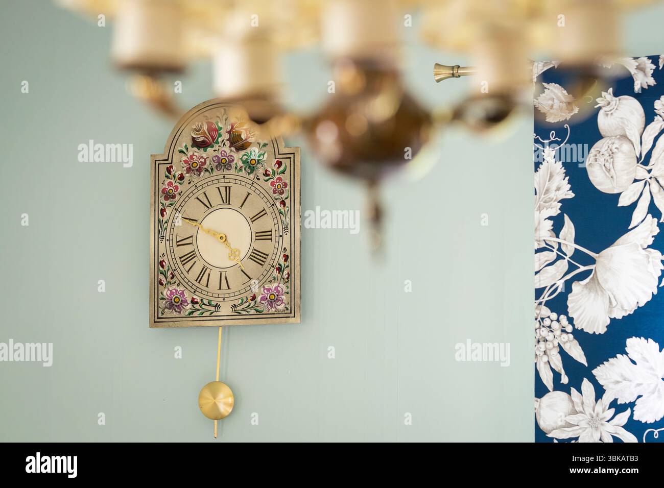Klassische hölzerne Wanduhr vor Tapetenhintergrund. Vor einem hellgrünen Hintergrund, eine klassische bemalte Wanduhr. Blaues Blumenmuster Stockfoto