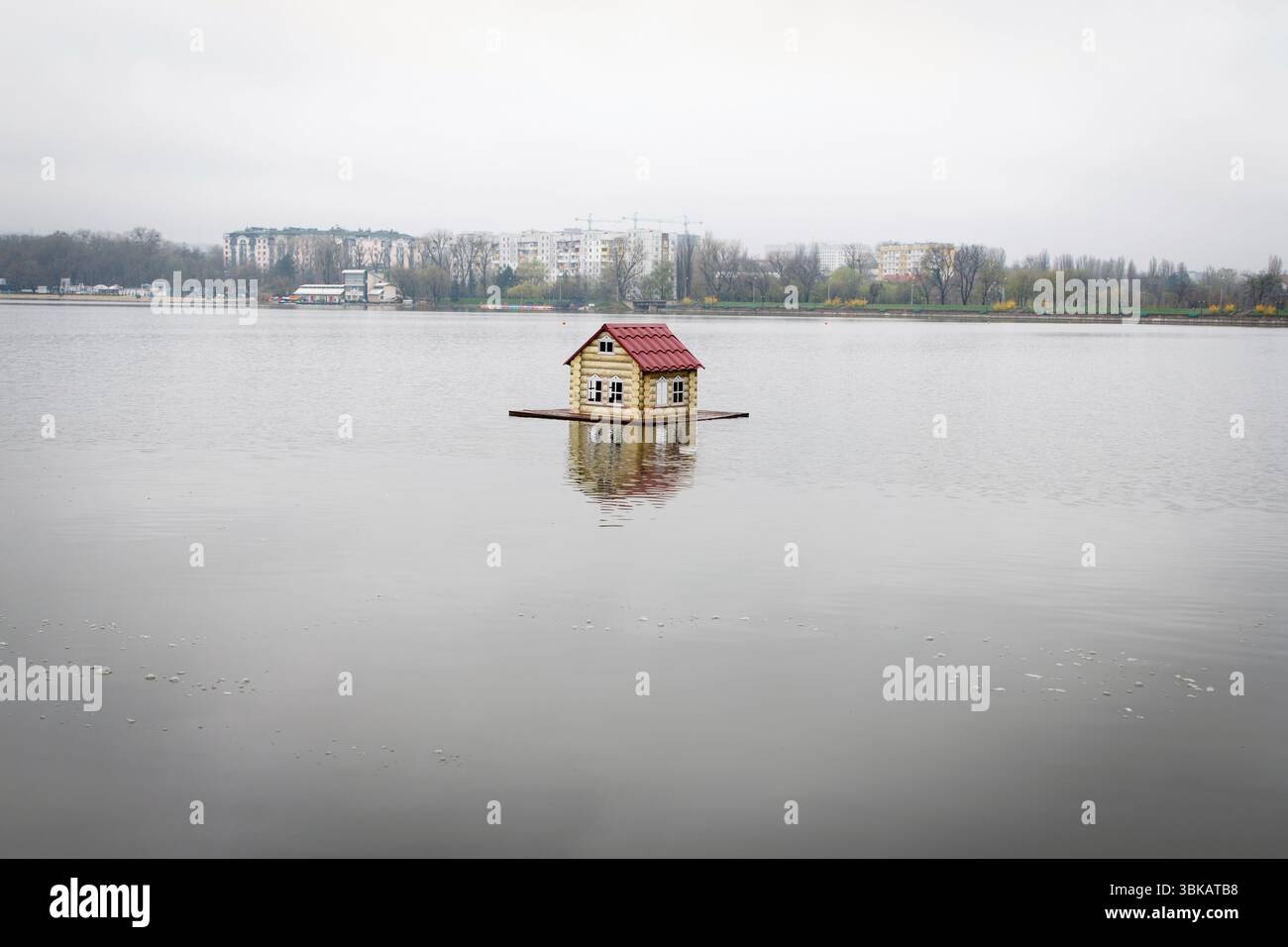 Winziges Holzhaus für Vögel mit rotem Dach, das auf einem ruhigen See schwimmt, bedeckter Himmel, Blick auf die ferne Stadt, Konzept der Isolation und urbaner Kontrast Stockfoto