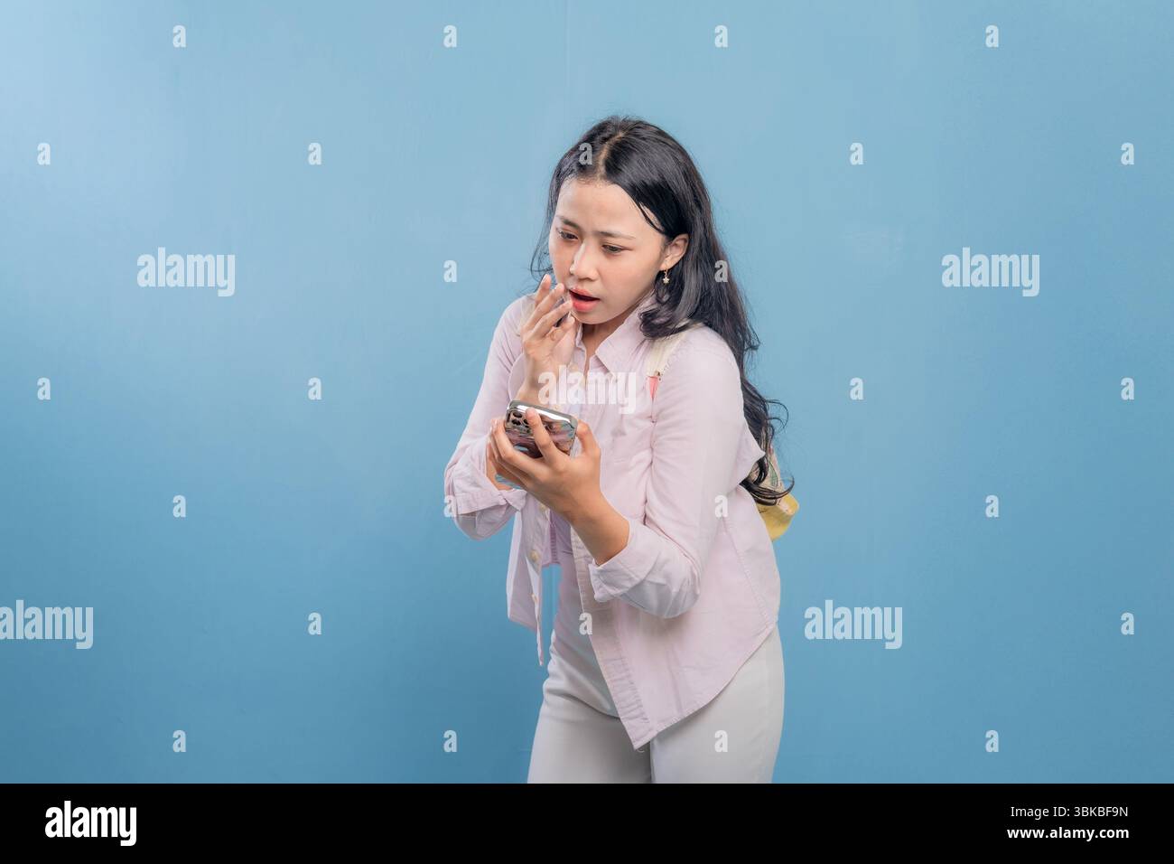 Eine Frau schaut auf ihr Handy, während sie vor einer blauen Wand steht. Sie ist von etwas auf ihrem Handy überrascht oder schockiert Stockfoto