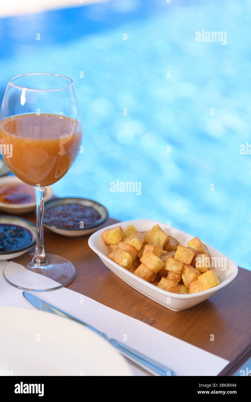 In Scheiben geschnittene Apfelwürfel, Pfirsichsaft und Marmelade auf einem Holztisch mit Blick auf den Pool im Hintergrund bei einem Hotelfrühstück. Stockfoto