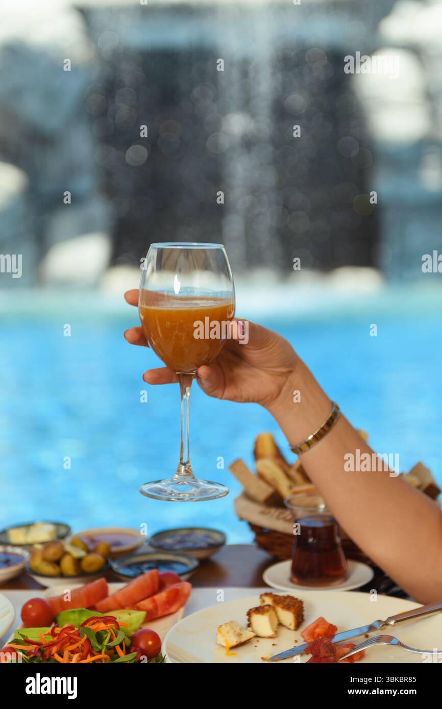 Ein Holztisch zum Frühstück im Hotel mit Salat, Oliven, Tomaten und Käse, während die Hand einer Frau ein Glas Saft hält. Es gibt einen ruhigen Pool Stockfoto