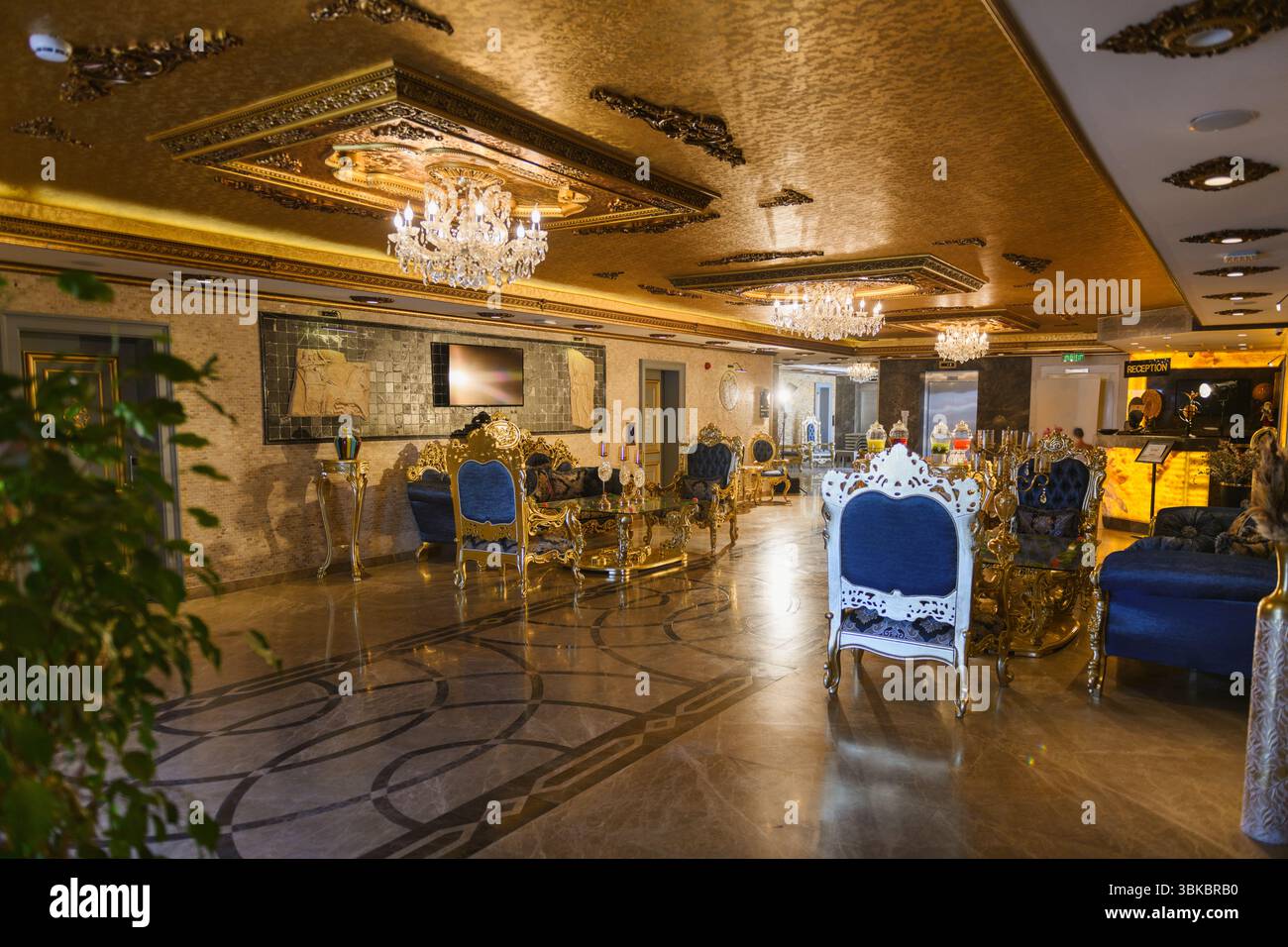 Opulente Hotellobby im Barockstil mit goldenen Akzenten, blauen Samtsesseln und Sofas, Marmorfußboden, großen Kronleuchtern und an der Wand montiertem Fernseher Stockfoto