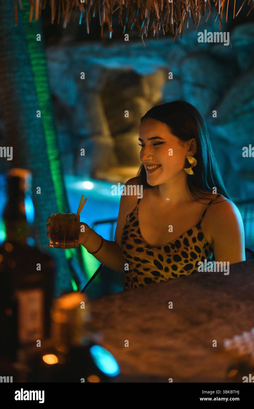 Stilvolle Brünette Frau im Leopardenkleid, die Getränke in der eleganten Hotelbar mit Pool im Hintergrund genießt. Stockfoto