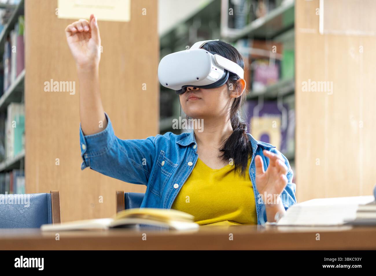 Eine junge Frau, die ein VR-Headset trägt, interagiert während des Studiums in einer Bibliothek mit virtuellen Inhalten und symbolisiert moderne Bildung und immersive Technologie. Stockfoto