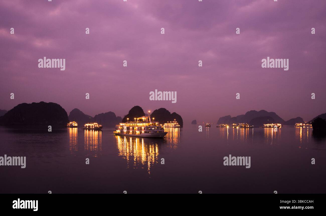 Boote mit leuchtenden Lichtern spiegeln sich in der Ha Long Bay bei Sonnenuntergang unter tiefviolettem Himmel Stockfoto