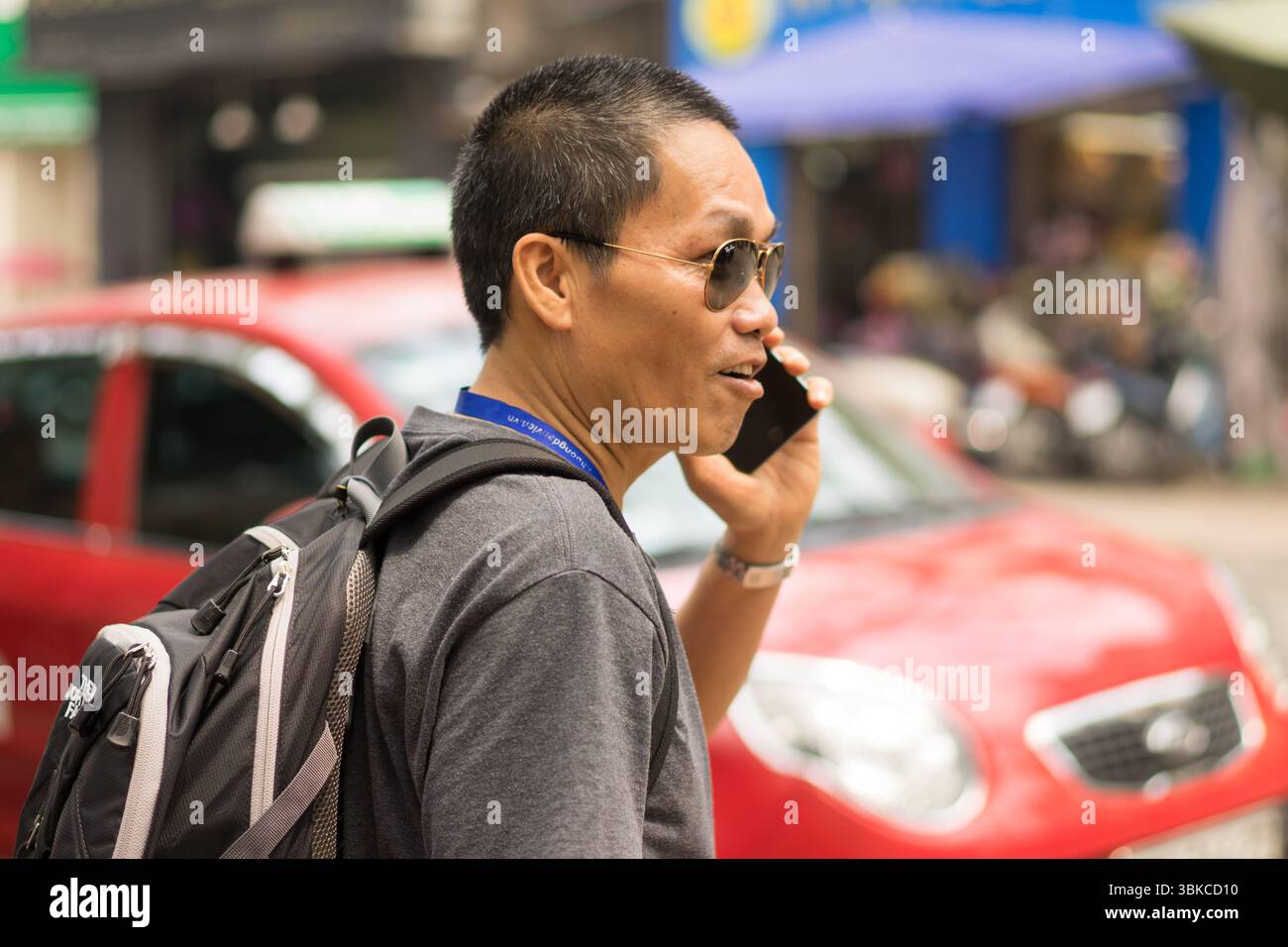 Vietnamesischer Mann mit kurzen dunklen Haaren in Schatten mit Rucksack am Telefon auf der Straße. Stockfoto
