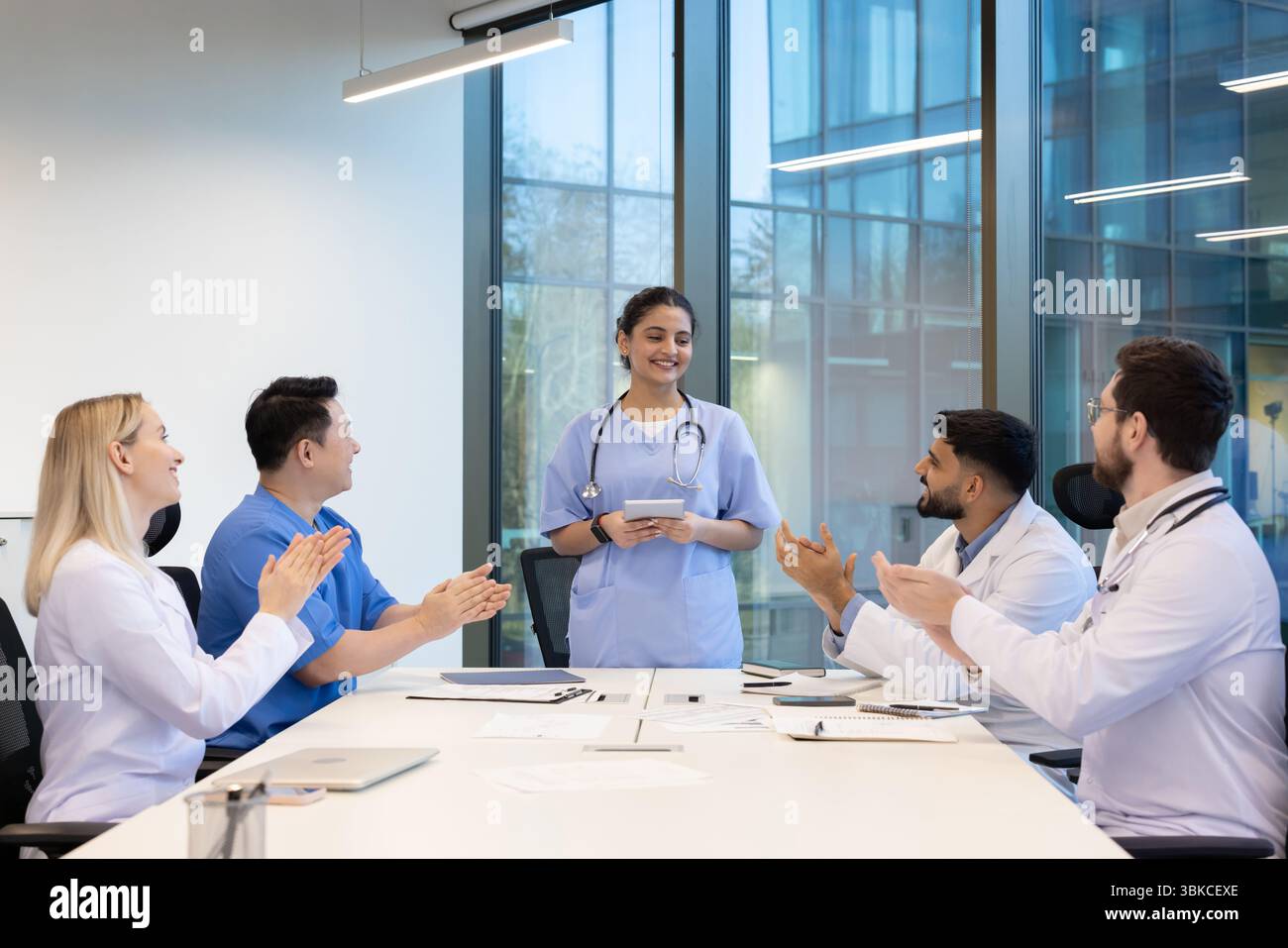 Ein vielfältiges medizinisches Team applaudiert in einem modernen Büroumfeld. Eine Ärztin steht, hält eine Tafel und lächelt. Stockfoto
