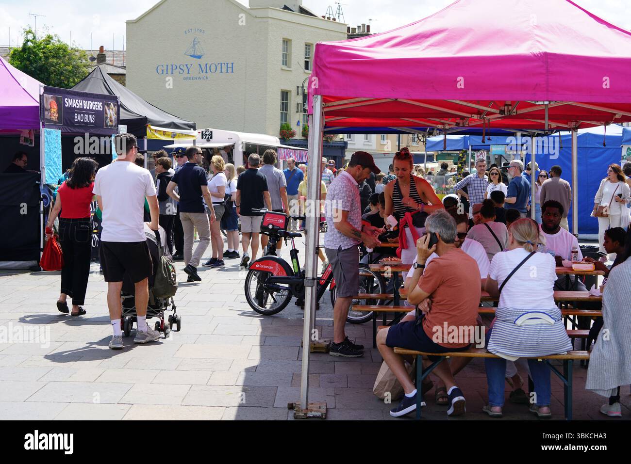 Geschäftige Marktszene im Freien mit Ständen, Tischen und Besuchern bei hellem Tageslicht. Greenwich, London, England Stockfoto