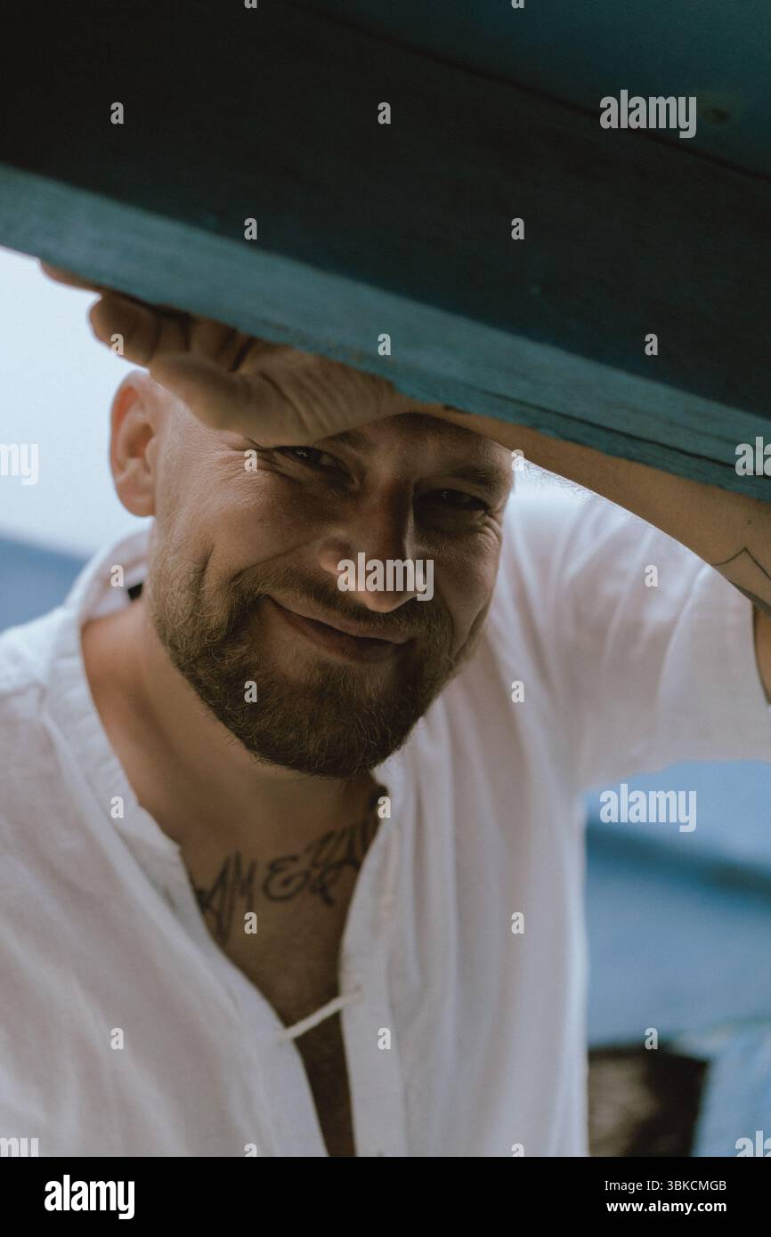 Lächelnder bärtiger Mann in weißem Hemd, der sich unter blauen Holzbalken beugt Stockfoto