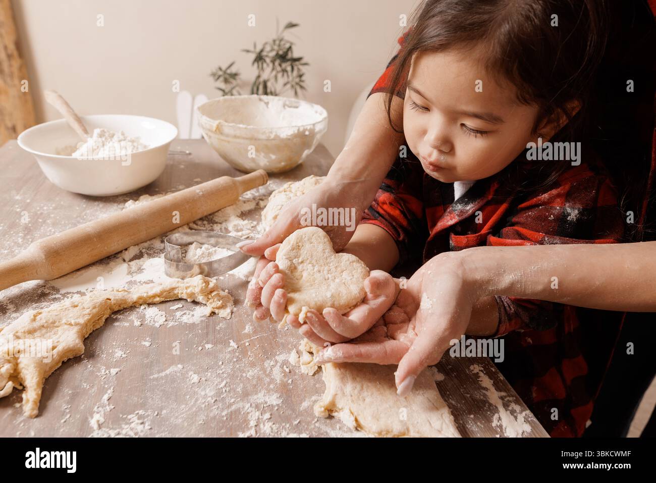 Kind und Mutter backen zusammen, halten herzförmigen Teig und symbolisieren Familienliebe, Sorgfalt und Freude in einer gemütlichen Küche. Stockfoto