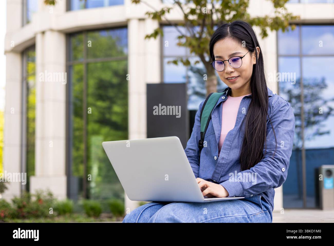 Eine junge Asiatin benutzt draußen einen Laptop, mit einem Gebäude und Grün im Hintergrund. Stockfoto