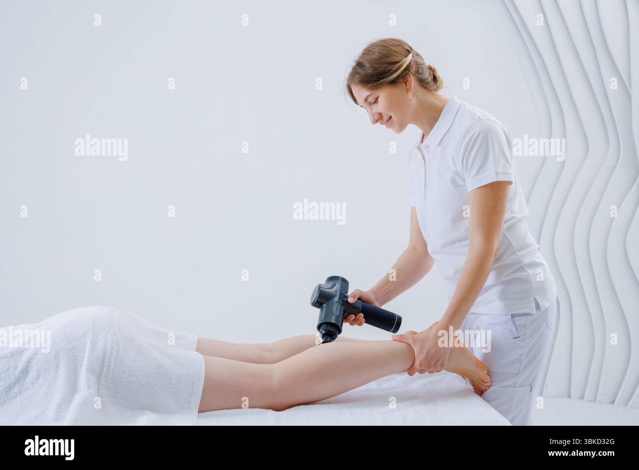 Professionelle Therapeutin, die Massagepistole Therapie auf den Unterschenkeln der Klienten anwendet, um Schmerzen zu lindern, den Blutfluss zu verbessern und die Erholung zu fördern. Stockfoto Professionelle Therapeutin, die Massagepistole Therapie auf den Unterschenkeln der Klienten anwendet, um Schmerzen zu lindern, den Blutfluss zu verbessern und die Erholung zu fördern. Stockfoto